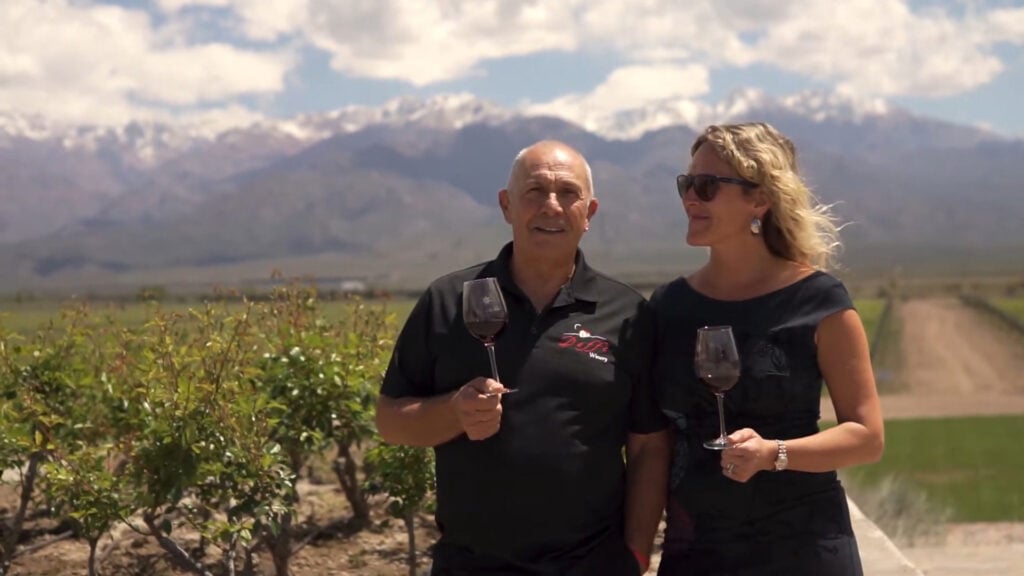 Two guests standing in a vineyard holding wine glasses, with the Andes Mountains in the background.